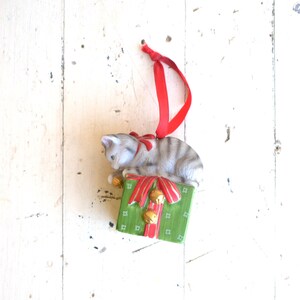 May include: A ceramic Christmas ornament featuring a gray tabby cat perched atop a green gift box. The box is decorated with a red ribbon and gold bells. A red ribbon is attached for hanging.