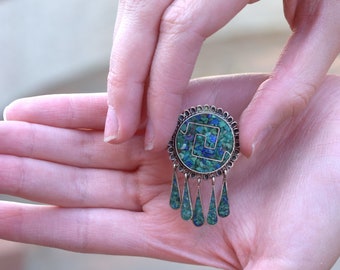 May include: A silver pendant with a turquoise and green stone design. The pendant has a geometric pattern and is hanging from a silver chain.