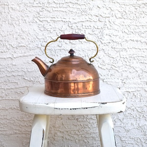 May include: A copper kettle with a dark wooden handle and brass accents. The kettle sits on a small, white, distressed stool. The kettle has a rounded body and a spout for pouring.