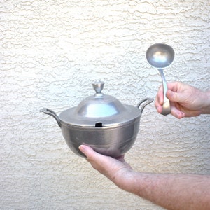 May include: A silver serving bowl with a lid and a matching ladle. The bowl has a round shape and a smooth surface. The ladle has a long handle and a round bowl.