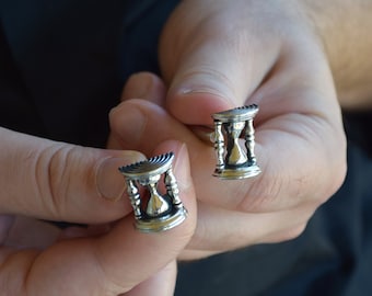 May include: Silver-toned cufflinks in the shape of hourglasses. The cufflinks are held in a person's hands, showcasing the intricate details of the design. The hourglasses have a detailed, vintage aesthetic.
