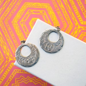May include: A pair of silver earrings with a circular design and intricate details. The earrings have a large, open circle in the center and are suspended from a small loop.