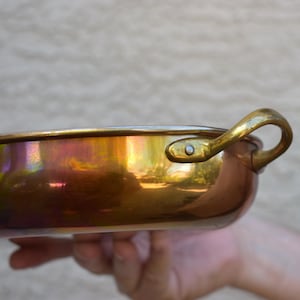 May include: A polished copper pan with a smooth, reflective surface showing iridescent highlights. The pan features a curved brass handle securely attached to the side with a single visible metal rivet, creating a metallic contrast.