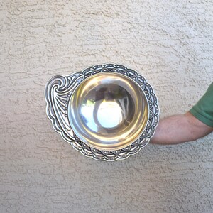 May include: A silver serving bowl with an ornate design around the rim and a decorative handle. The bowl is made of metal and has a shiny finish.