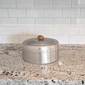 May include: A round, silver-colored metal cake carrier with a wooden knob on the lid. The base of the carrier has a decorative floral pattern. The carrier is sitting on a speckled countertop in front of a white tile backsplash.