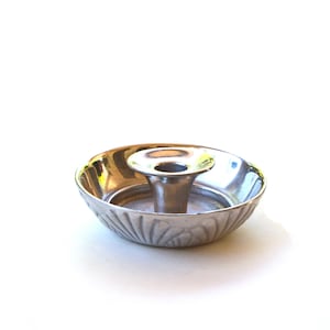 May include: A small, silver-colored candle holder with a central cup and a decorative, ridged bowl. The candle holder is made of metal and sits on a white surface. The bowl has a leaf-like pattern.