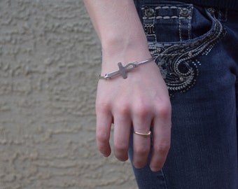 May include: A silver bracelet with an ankh charm. The bracelet is on a person's wrist, and they are also wearing a gold ring on their finger.
