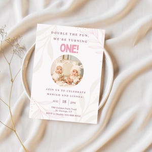 May include: A pink and white invitation card for a first birthday celebration. The card features the text "DOUBLE THE FUN, WE'RE TURNING ONE!" and includes a photo of two babies wearing bunny hats. The invitation also includes the date, time, and location.