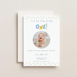 Op de afbeelding: Een witte uitnodigingskaart met de tekst "IT'S SO FUN TO BE ONE!" in kleurrijke letters. Een foto van een lachende baby staat in een cirkel. De kaart bevat ook evenementdetails en RSVP-informatie.