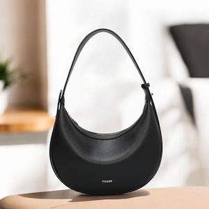 May include: Black crescent-shaped handbag with a single shoulder strap. The bag has a smooth texture and a zipper closure. The brand name "FOXER" is printed in small letters on the front of the bag. The bag is sitting on a light brown surface.