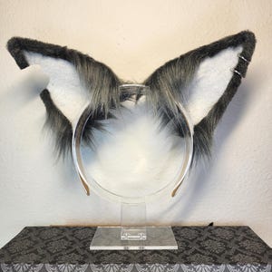 May include: Gray furry cat ears headband with a white inner lining. The headband is made of clear plastic and is on a white stand.