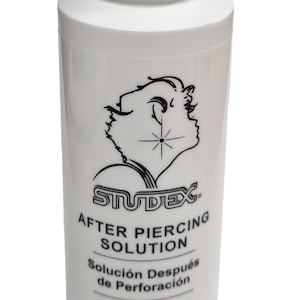 May include: White bottle of Studex After Piercing Solution. The bottle has a white flip-top cap and a black and white graphic of a profile with a starburst. The bottle contains 4 fl oz / 100ml of solution.
