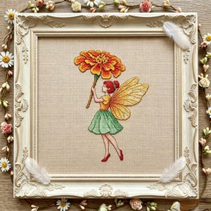 Könnte beinhalten: Eine Kreuzstichstickerei mit einer Fee mit roten Haaren, gelben Flügeln, einem gelben Oberteil und einem grünen Rock, die eine große orange-gelbe Studentenblume hält. Das Kunstwerk befindet sich in einem verzierten, cremefarbenen Rahmen.