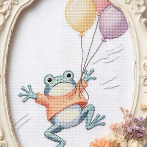 Könnte beinhalten: Kreuzstich-Kunstwerk einer Froschfigur, die drei Ballons in einem dekorativen, verzierten, cremefarbenen Rahmen hält. Der Frosch ist hellblau mit einem orangefarbenen Hemd. Die Ballons sind gelb, rosa und lila. Das Kunstwerk ist mit Trockenblumen verziert.