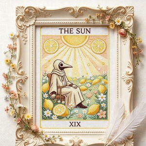 Könnte beinhalten: Kreuzstich-Tarotkartenkunst mit dem Titel "THE SUN", die eine Figur mit einer Pestdoktormaske zeigt, die in einem Feld aus Zitronen und Blumen sitzt. Das Kunstwerk ist in einem dekorativen weißen Rahmen gerahmt.