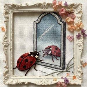 Puede incluir: Una obra de arte en punto de cruz enmarcada que presenta una mariquita roja con manchas negras, frente a su reflejo en un espejo. El espejo tiene un fondo azul. El marco es blanco con detalles ornamentados, y la obra está decorada con pequeñas flores secas.