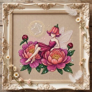 Könnte beinhalten: Eine Kreuzstichstickerei in einem cremefarbenen, verzierten Rahmen mit einer Fee in einem rosa Blütenhut und Kleid, die auf großen rosa Pfingstrosen sitzt. Über der Fee ist ein weißes Waage-Sternbild gestickt, ergänzt durch weiße Federn.