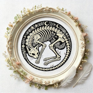 Peut inclure: Une broderie circulaire représentant un squelette de chat couleur crème sur fond noir, encadrée dans un cadre ovale blanc cassé orné de petites fleurs pâles et de deux plumes blanches placées dans le coin inférieur droit.