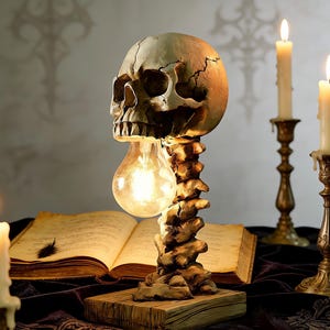 Könnte beinhalten: Eine schädelartige Lampe mit einer Glühbirne anstelle des Gehirns, getragen von einem wirbelsäulenförmigen Ständer. Die Lampe steht auf einem Holzsockel, mit einem offenen Buch und brennenden Kerzen im Hintergrund. Vintage, gotische Ästhetik.