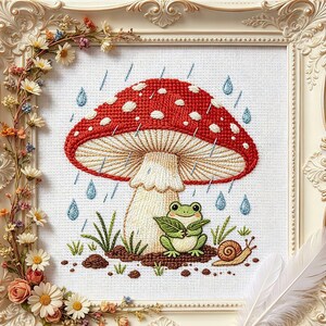 Könnte beinhalten: Eine Kreuzstichstickerei mit einem großen roten Pilz mit weißen Punkten, einem kleinen grünen Frosch, der ein Blatt hält, und einer braunen Schnecke. Die Szene zeigt blaue Regentropfen und grünes Gras, eingefasst in einen cremefarbenen Rahmen.