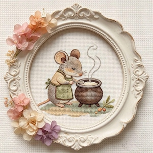 Könnte beinhalten: Kreuzstich-Stickerei einer Maus mit Schürze, die in einem Topf rührt. Das Kunstwerk befindet sich in einem weißen, verzierten, ovalen Rahmen, der mit getrockneten Blumen in Rosa- und Pfirsichtönen verziert ist. Dampf steigt aus dem Topf auf.