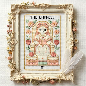 May include: Cross-stitch artwork of "The Empress" tarot card, framed in a cream-colored ornate frame with floral accents. The card features a woman in a pink dress, surrounded by flowers and strawberries. The Roman numeral III is at the bottom.