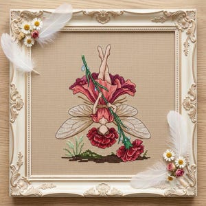 May include: A cross-stitch embroidery of a fairy hanging upside down from a green stem, wearing a dark pink petal dress with translucent wings, framed in an ornate cream-colored frame decorated with white feathers and small daisies.