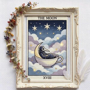 Könnte beinhalten: Kreuzstich-Kunstwerk in einem cremefarbenen, verzierten Rahmen. Das Bild zeigt eine Pestdoktorfigur in einer halbmondförmigen Tasse, vor einem sternenklaren Nachthimmel und flauschigen Wolken. Der Text "THE MOON" und "XVIII" sind sichtbar.