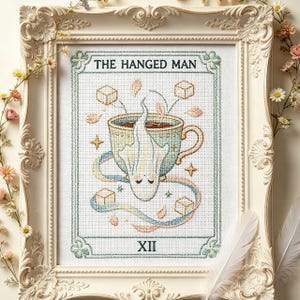 Könnte beinhalten: Gerahmtes Kreuzstich-Kunstwerk mit einem Tarotkarten-Design. Das Kunstwerk zeigt eine Teetasse mit einer geisterhaften Figur, Würfeln und den Texten "THE HANGED MAN" und "XII". Der Rahmen ist verziert, cremefarben und mit floralen Akzenten versehen.