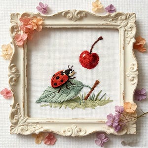 Könnte beinhalten: Gesticktes Bild in einem weißen, verzierten Rahmen. Es zeigt einen roten Marienkäfer mit schwarzen Punkten auf einem grünen Blatt, eine rote Kirsche und Gras. Getrocknete rosa und lila Blumen umgeben den Rahmen.