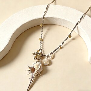 Op de afbeelding: Een lange, antiek goudkleurige ketting met een grote, stekelige schelpenhanger. De ketting bevat een gouden bloemcharm, kleine gouden schijfjes en schelp- en parelaccenten. De ketting is delicaat en het ontwerp roept een strandachtige, bohemien esthetiek op.