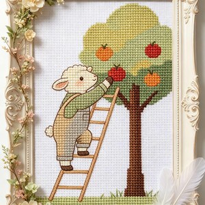 Könnte beinhalten: Kreuzstich-Kunstwerk eines Schafs in Latzhosen auf einer Leiter, das Äpfel von einem Baum pflückt. Der Baum hat rote und orangefarbene Äpfel. Das Kunstwerk befindet sich in einem weißen, verzierten Rahmen mit floralen Akzenten.