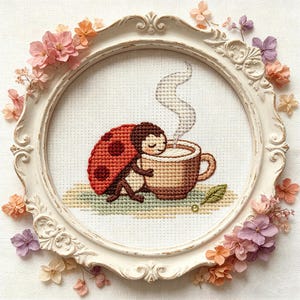 Könnte beinhalten: Ein gerahmtes Kreuzstich-Kunstwerk zeigt einen Marienkäfer, der eine Kaffeetasse umarmt, aus der Dampf aufsteigt. Der Marienkäfer hat einen rot-schwarz gepunkteten Panzer. Das Kunstwerk befindet sich in einem weißen, verzierten Rahmen, der mit pastellfarbenen Blumen verziert ist.