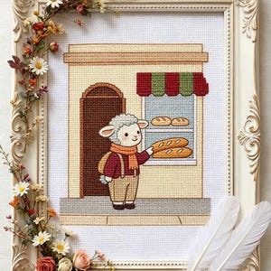 Könnte beinhalten: Kreuzstich-Kunst eines Schafcharakters, der ein Brot vor einer Bäckerei hält. Das Kunstwerk ist in einem weißen, verzierten Rahmen mit floralen Akzenten gerahmt. Die Bäckerei hat eine gestreifte Markise und eine braune Tür.