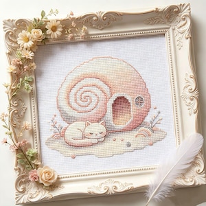 Könnte beinhalten: Eine Kreuzstichstickerei mit einer schlafenden weißen Katze vor einem großen, spiralförmigen rosa Schneckenhaus. Das Kunstwerk befindet sich in einem verzierten, cremefarbenen Rahmen, der mit kleinen Stoffblumen und einer weißen Feder geschmückt ist.