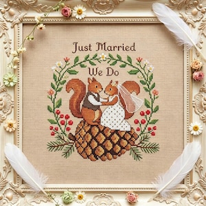 Könnte beinhalten: Eine Kreuzstichstickerei mit zwei Eichhörnchen auf einem Tannenzapfen, eines trägt eine Weste, das andere ein weißes Kleid mit Schleier. Der Text Just Married We Do ist von einem Kranz aus grünen Blättern, roten Beeren und weißen Blumen umgeben, alles in einem cremefarbenen Rahmen mit weißen Federn und kleinen Blüten.