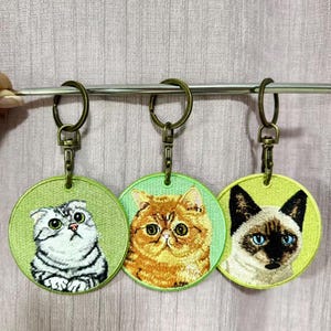 May include: Three embroidered cat keychains. Each keychain features a different cat breed: a gray and white Scottish Fold, an orange Exotic Shorthair, and a Siamese cat. The keychains have a green background and a bronze-colored metal ring.