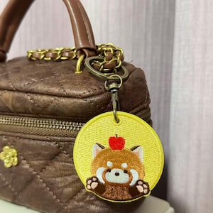 Puede incluir: Un bolso acolchado marrón con una correa de cadena dorada y un asa. Un llavero circular amarillo con un diseño de panda rojo y una manzana roja en la cabeza está sujeto a la bolsa.