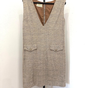 Op de afbeelding: Een mouwloze beige jurk met een donkerbruin ruitpatroon, een diepe V-hals en twee voorzakken met kleppen en goudkleurige knopen. De binnenkant is gevoerd met bruine stof en een label met de tekst Butterick is zichtbaar bij de halslijn.