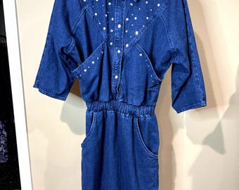 May include: A dark blue denim dress with a collared neckline and three-quarter sleeves. The dress features silver-colored embellishments and pockets. The waist is elasticized. The dress is hanging on a black hanger.