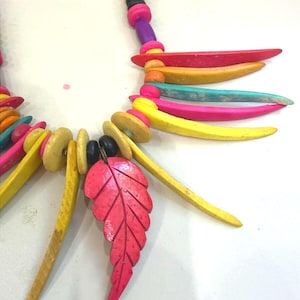 May include: A necklace featuring a vibrant array of multi-colored wooden beads and elongated, curved wooden spikes in shades of yellow, pink, orange, and teal. A prominent, carved pink leaf-shaped pendant hangs from the center of the strand.