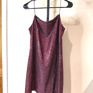 May include: A burgundy sequined slip dress hangs on a black hanger. The dress has thin spaghetti straps and a V-neckline. The fabric is covered in small, reflective sequins, creating a shimmering effect. The dress is knee-length and appears to be made of a lightweight material.