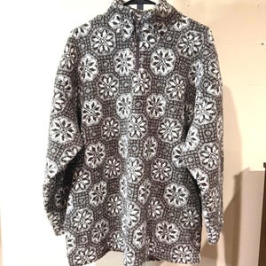 May include: A gray and white patterned fleece pullover with a half-zip collar. The sweater features a repeating snowflake design. The long-sleeved top is hanging on a black hanger.