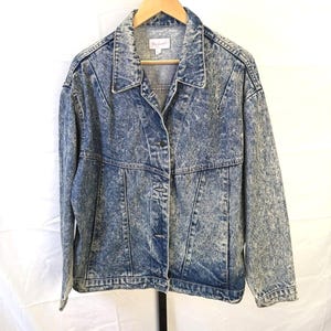 May include: Acid wash denim jacket with a button-up front and collared neckline. The jacket is hanging on a wooden hanger. The jacket has a light blue and white marbled pattern.