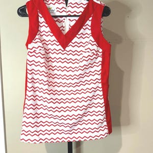 Op de afbeelding: Een mouwloze top met een witte basis en een herhalend rood chevronpatroon. Het kledingstuk heeft een effen rode V-hals en effen rode zijpanelen. Een witte rits is zichtbaar aan de achterkant en er is een klein label in de nek genaaid.