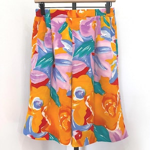 May include: A pair of colorful shorts with a vibrant floral pattern. The shorts feature a bright orange background with large flowers in shades of yellow, pink, purple, and blue. The shorts are hanging on a wooden hanger.