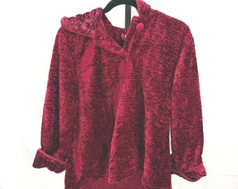 Puede incluir: Chaqueta burdeos texturizada con mangas largas y cierre de botón redondo. La chaqueta tiene un cuello único y está colgada de una percha negra sobre un fondo blanco. Las mangas tienen un puño decorativo.