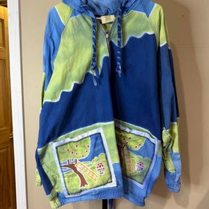 May include: A blue, green, and white hooded pullover with a half-zip closure. The pullover features a unique design with tree and mushroom graphics on the front pockets. The sleeves and hood are a lighter blue and green, while the body is a darker blue.