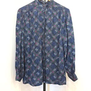 May include: A long-sleeved blue blouse with a high collar and button-down front. The fabric features a plaid pattern with white, pink, and green accents, and a repeating white floral design. The blouse is hanging on a wooden hanger.