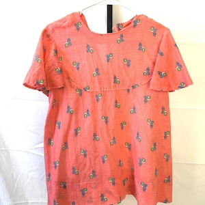 May include: Coral-colored top with short sleeves and a round neckline. The fabric is patterned with small blue, yellow, and white floral designs. The top is hanging on a white hanger against a white background.